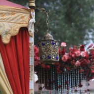Intricate brass lantern hanging near a red curtain with flowers in the background.