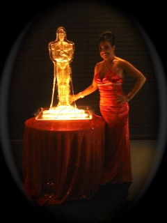 A woman in a red dress poses beside a glowing Oscar statue on a round table.