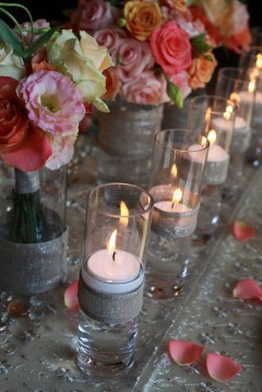 Row of lit candles and pink flowers in glass jars on a stone surface.
