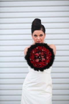 Brunette woman in white dress holding a bouquet of red roses with black feathers.