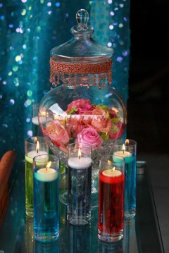 A decorative glass jar filled with pink roses, surrounded by colorful lit candles.