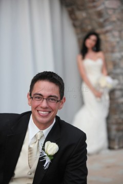 Smiling groom in glasses with bride blurred in the background.